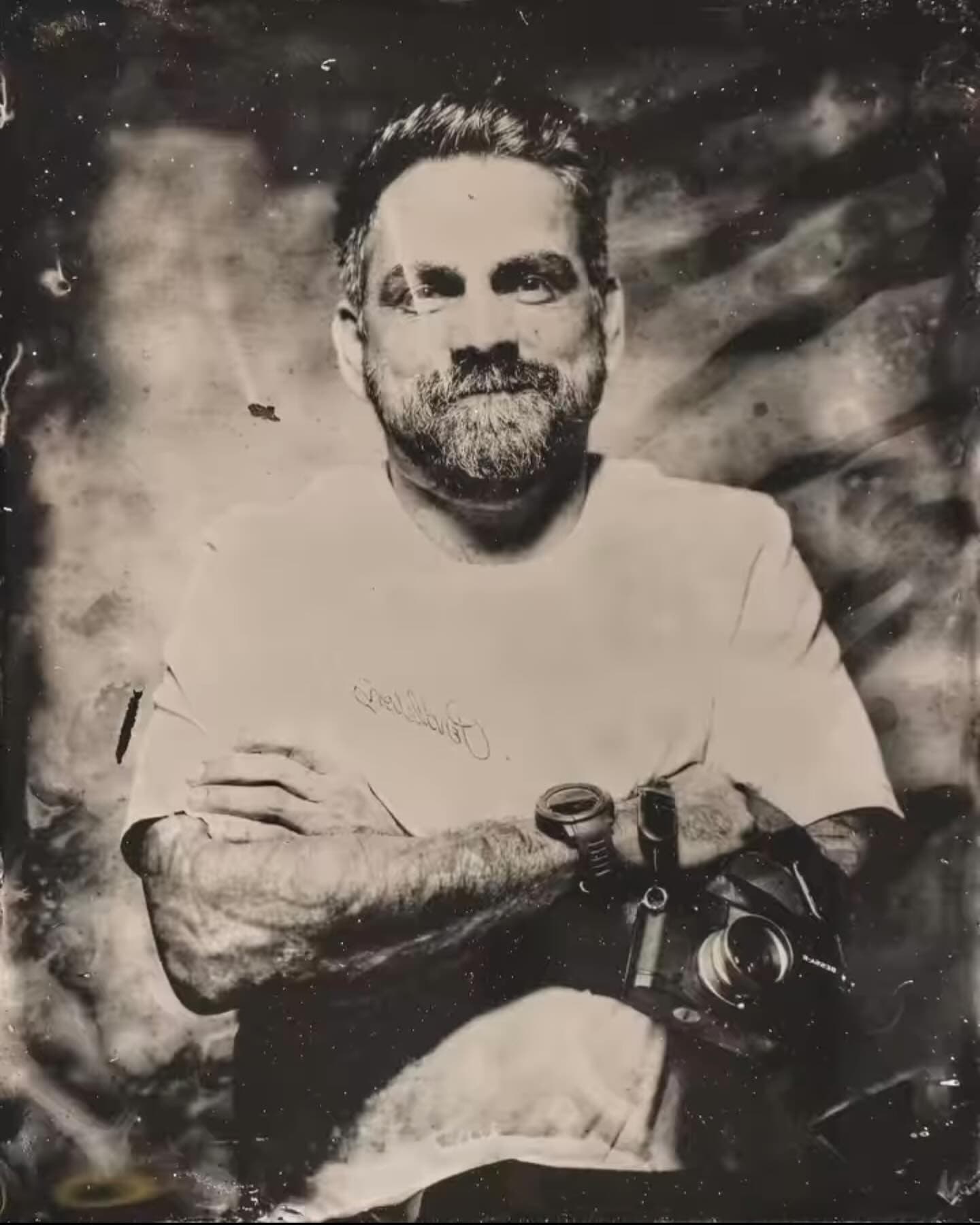 Black-and-white wet-plate portrait of Erick Florez, arms crossed, holding a film camera